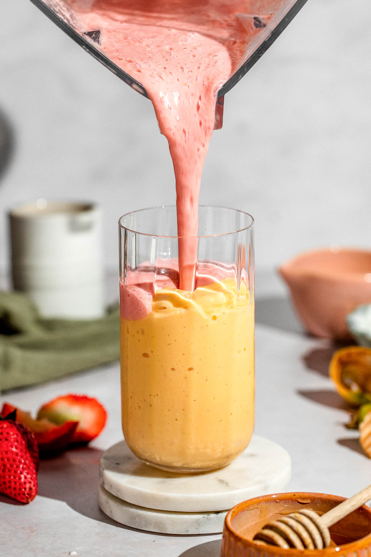 Strawberry smoothie being poured into a serving glass half filled with mango smoothie.