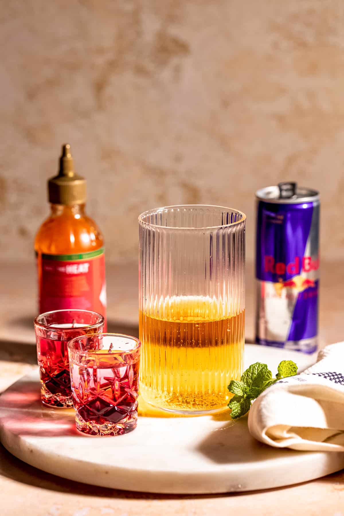 A tall glass half filled with Red Bull sitting next to two shot glasses with red liquid.