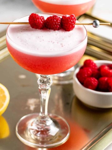 Two clover club cocktails served in coupe glasses sitting on a gold tray.