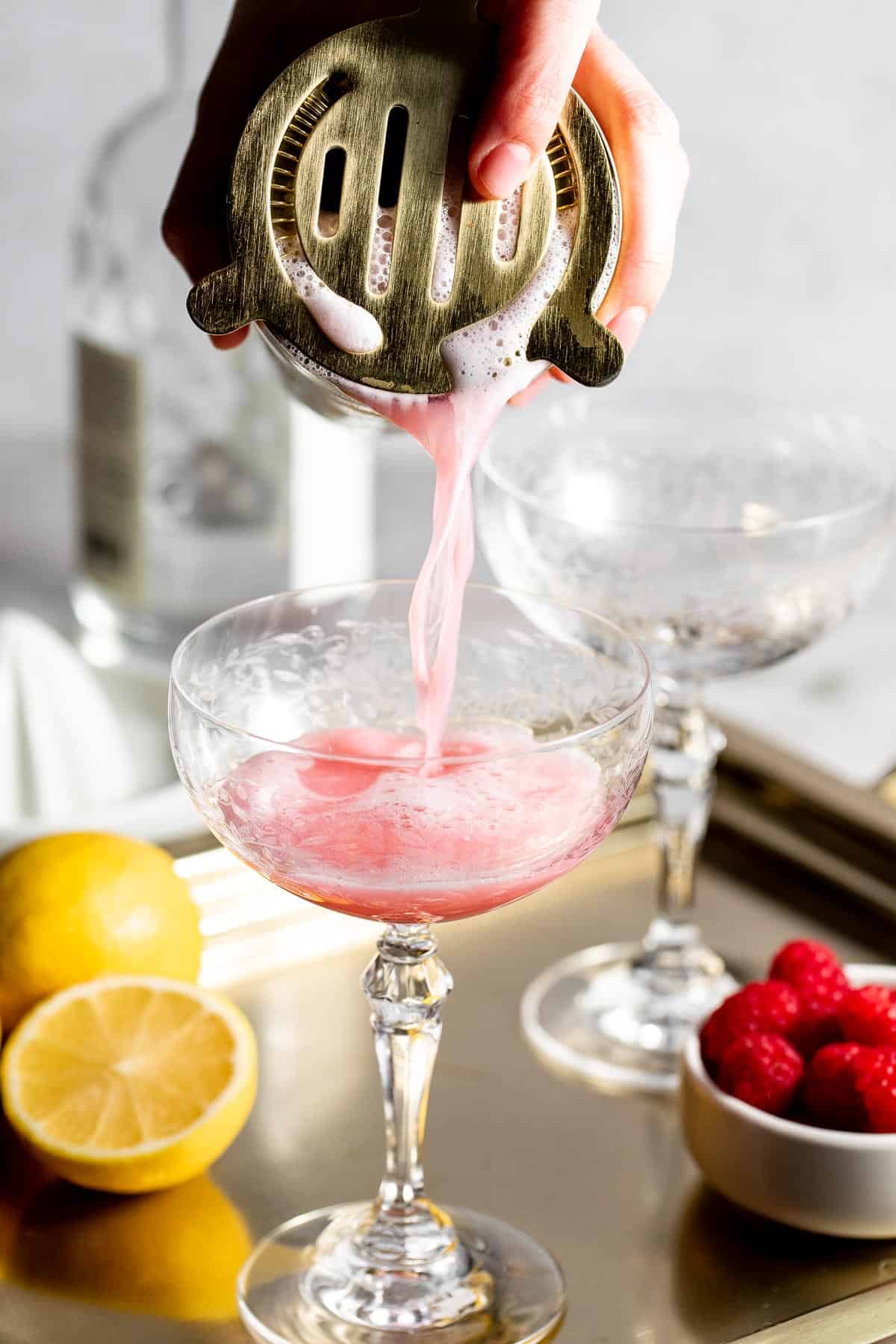 A foamy clover club cocktail being poured from a shaker through a cocktail strainer.