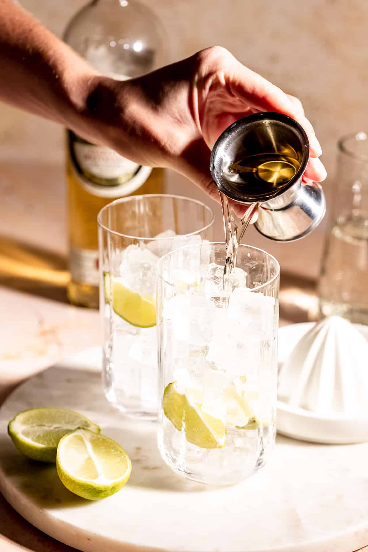 Tequila being poured into a highball glass with ice and lime wedges.