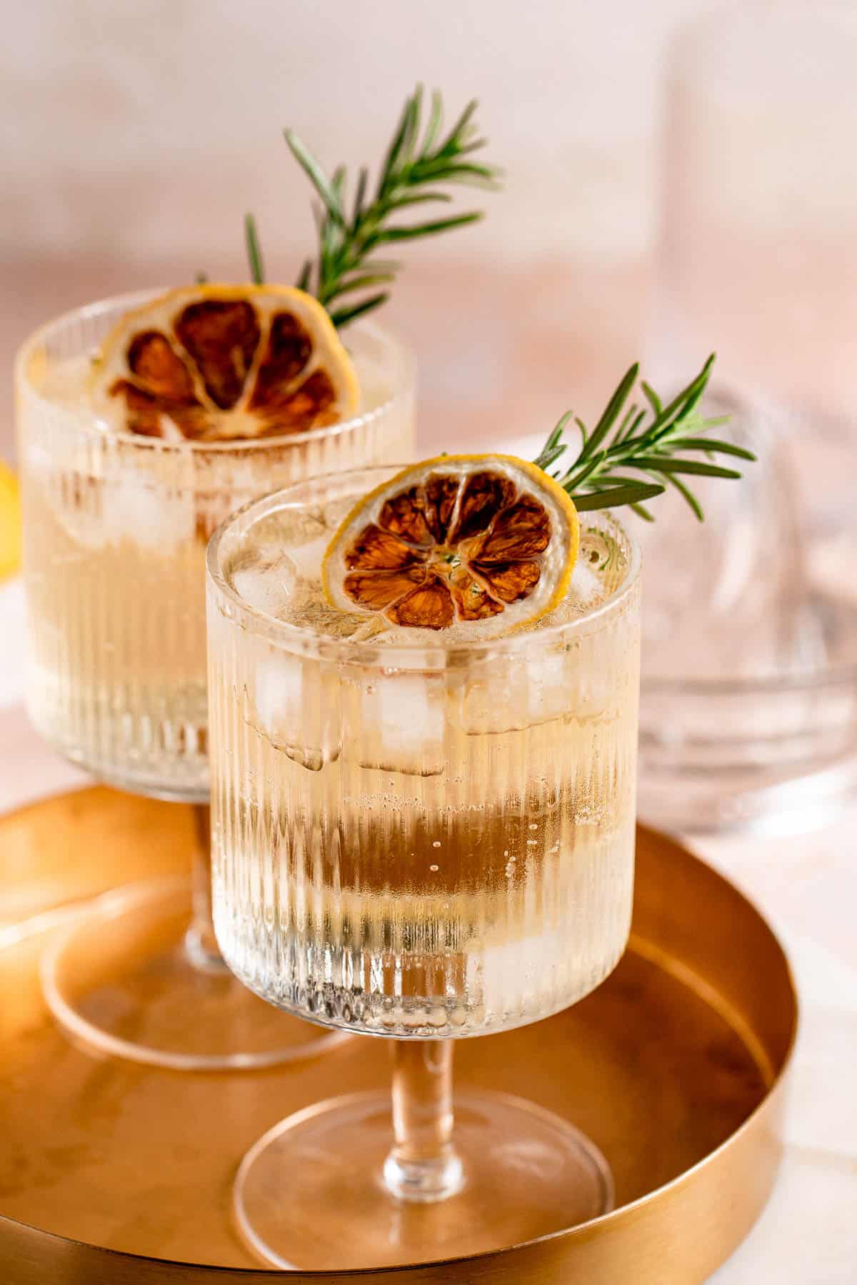 Two whiskey and tonics garnished with fresh herbs and lemon slices sitting on a gold tray.