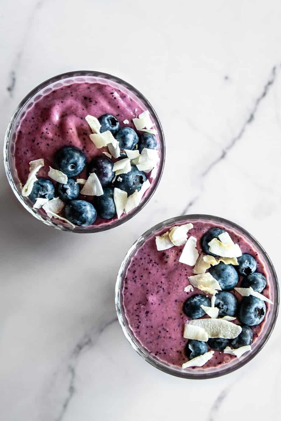 Two glasses of blueberry pineapple smoothie topped with coconut chips and fresh berries.