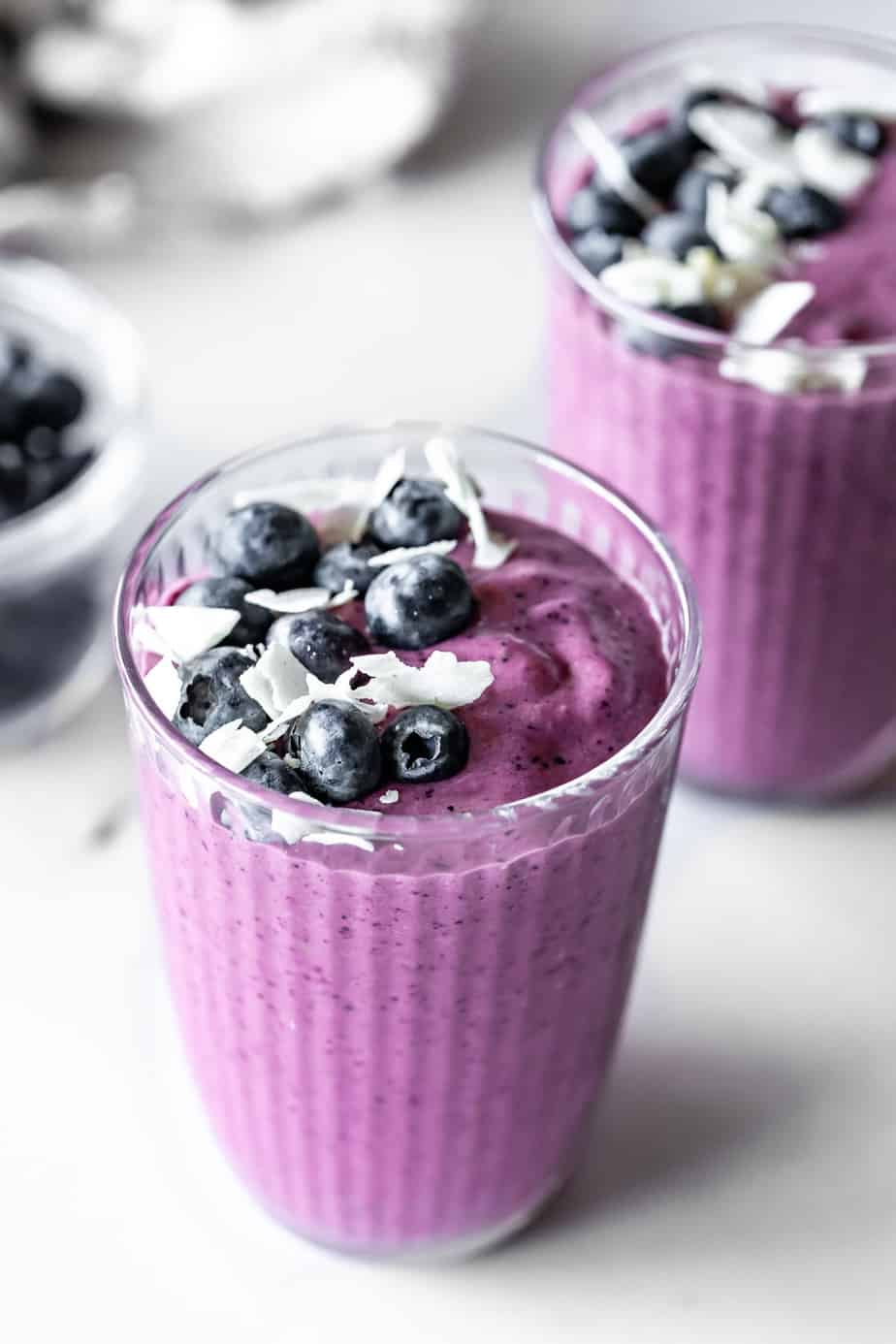 A tall glass filled with blueberry pineapple smoothie topped with coconut flakes and fresh blueberries.