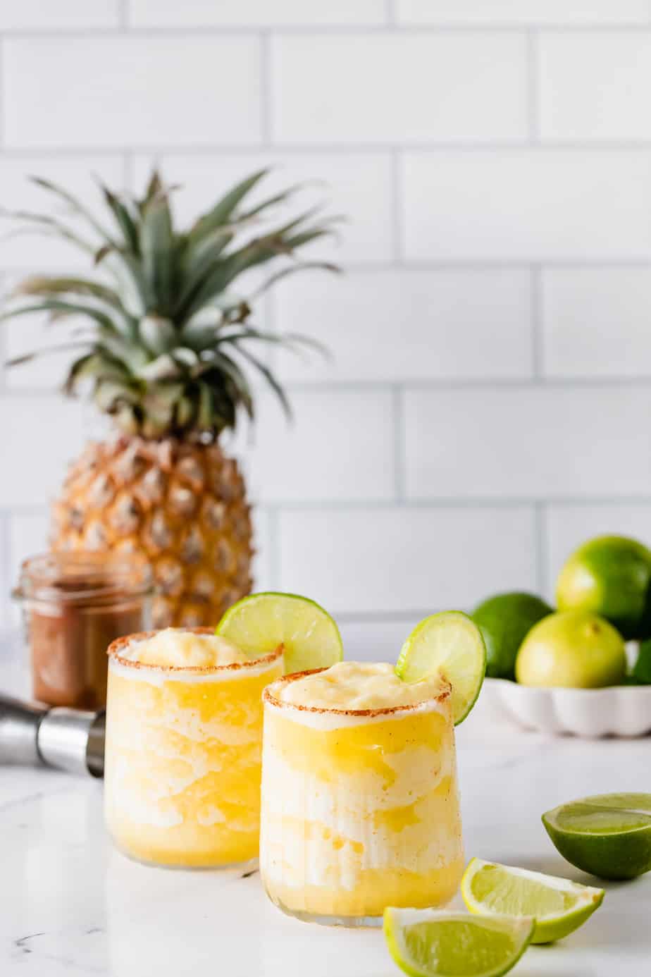 Two glasses filled with spicy pineapple margarita sitting on a countertop next to a fresh pineapple and limes.