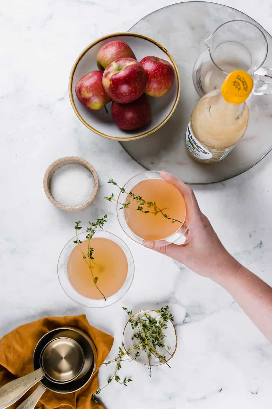 A hand holding a spiced apple cider cocktail garnished with fresh thyme.