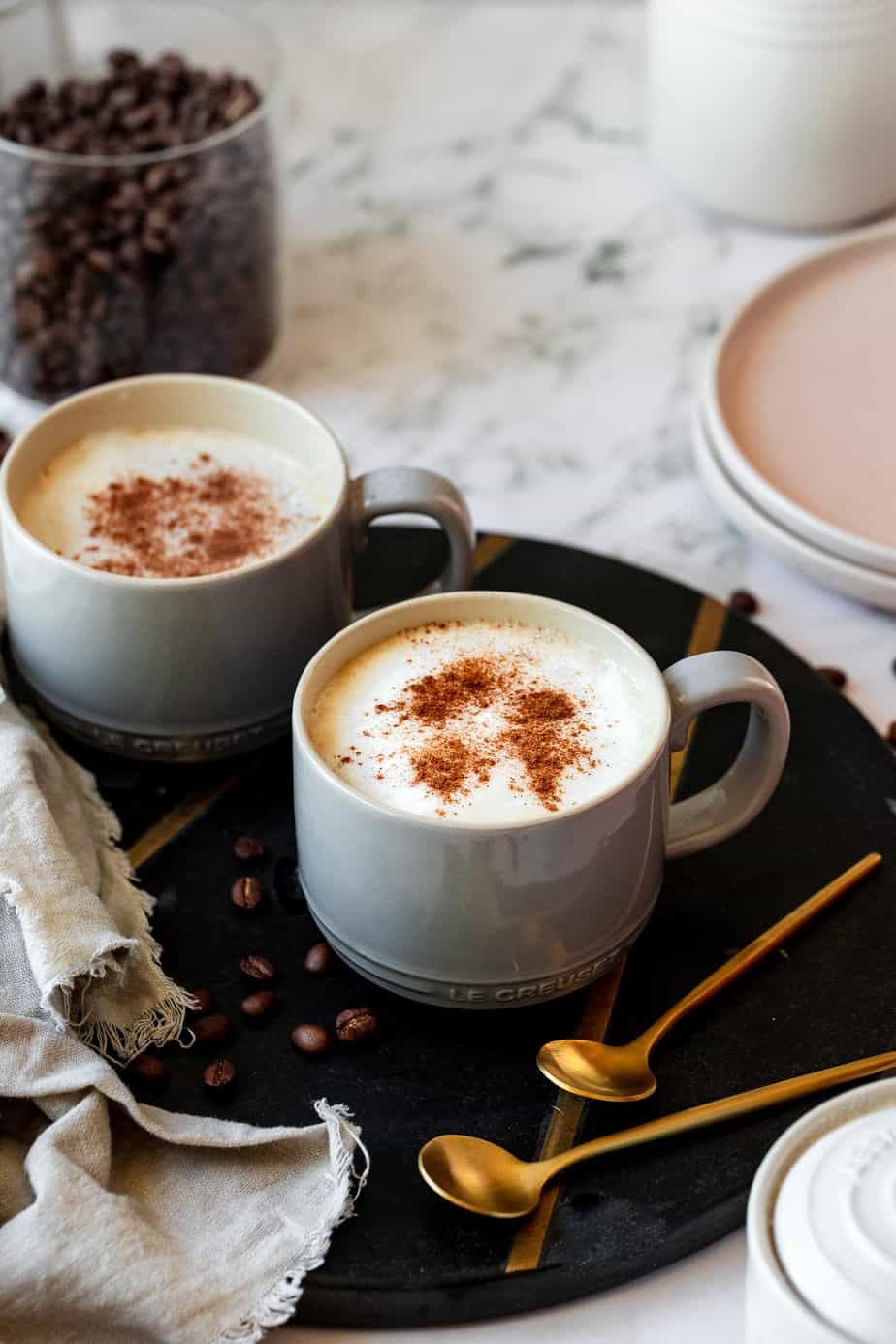 Two coffee mugs filled with oat milk lattes sitting on a serving tray with some coffee beans.