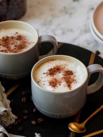 Two coffee mugs filled with oat milk lattes topped with ground cinnamon.