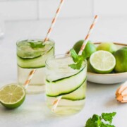 Two cucumber gin and tonics garnished with fresh mint and lime sitting on a countertop.