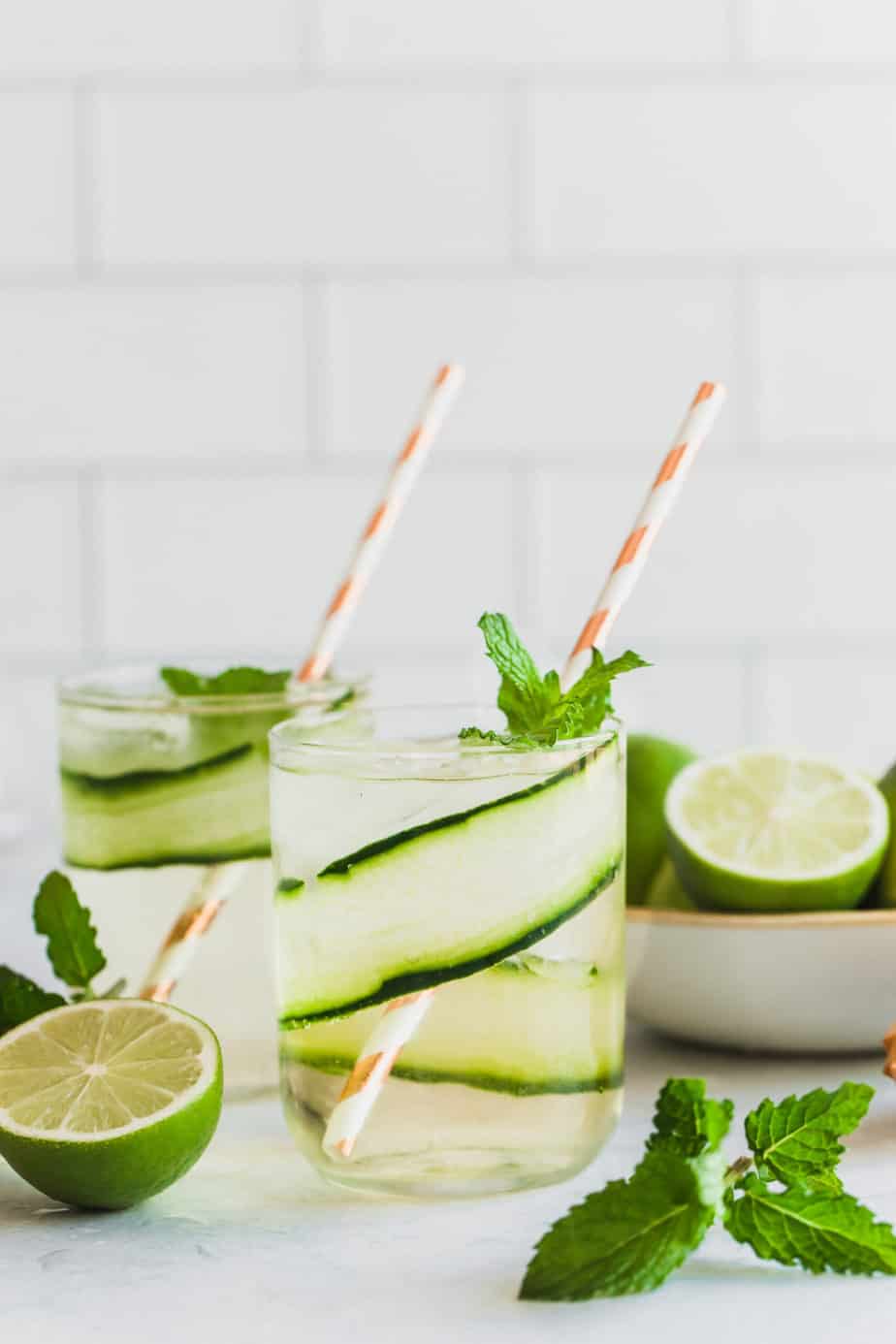 Two glasses of cucumber gin and tonic garnished with limes and mint leaves.