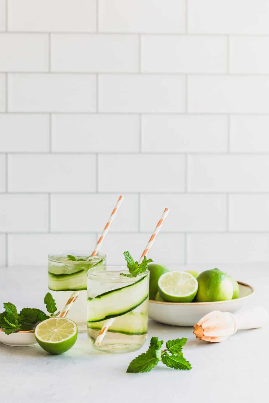 Two cucumber gin and tonic cocktails garnished with fresh mint and limes sitting on a kitchen counter.