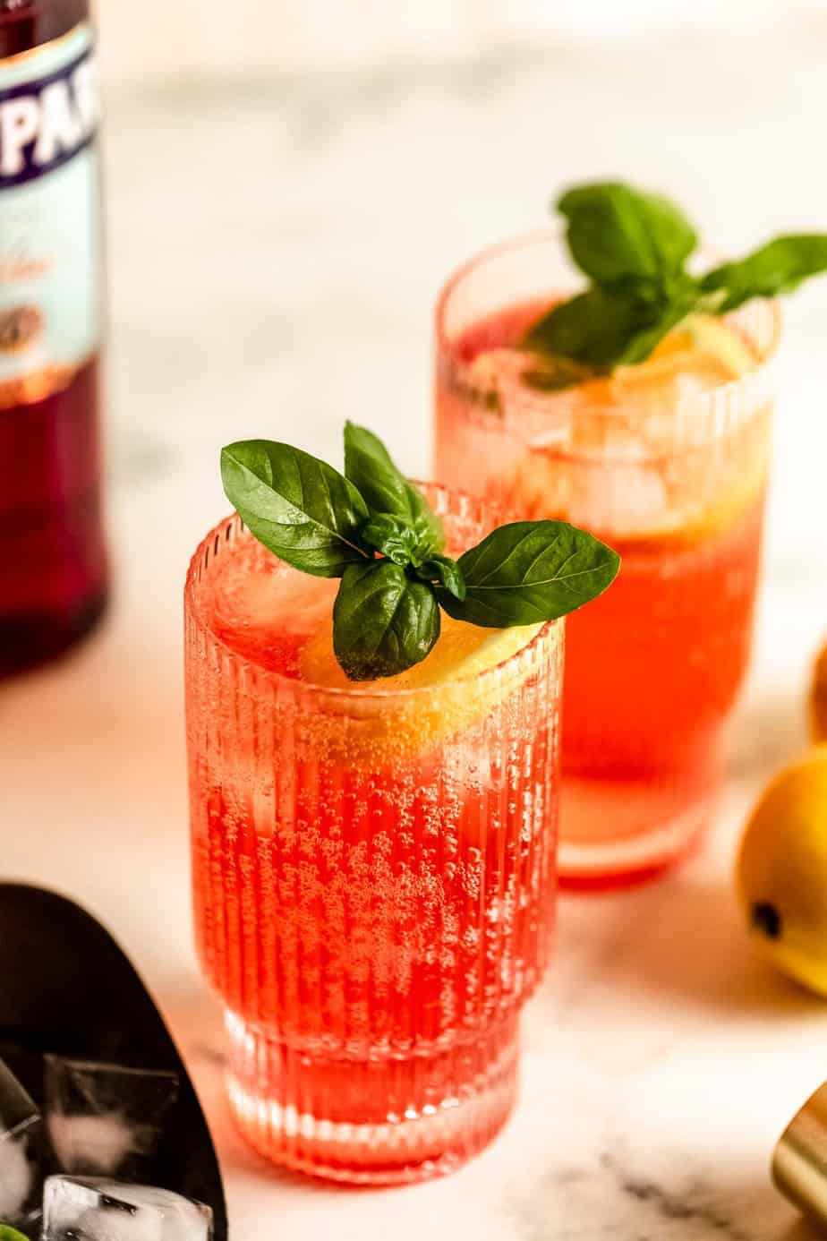 Two glasses of Campari and soda garnished with fresh basil and slices of lemon.