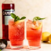 Two glasses of Campari soda garnished with fresh basil and lemon wedges sitting on a counter.