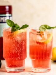 Two glasses of Campari soda garnished with fresh basil and lemon wedges sitting on a counter.