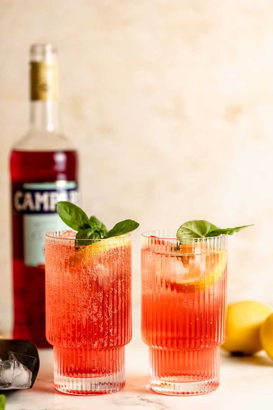 Two Campari and soda served with fresh basil and lemon slices sitting on a counter.