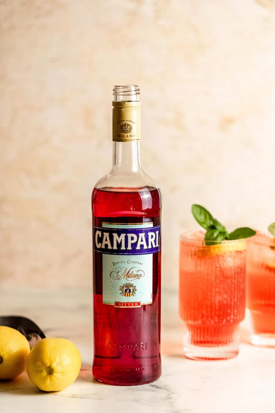 A bottle of Campari sitting on a counter next to two drinks and some lemons.