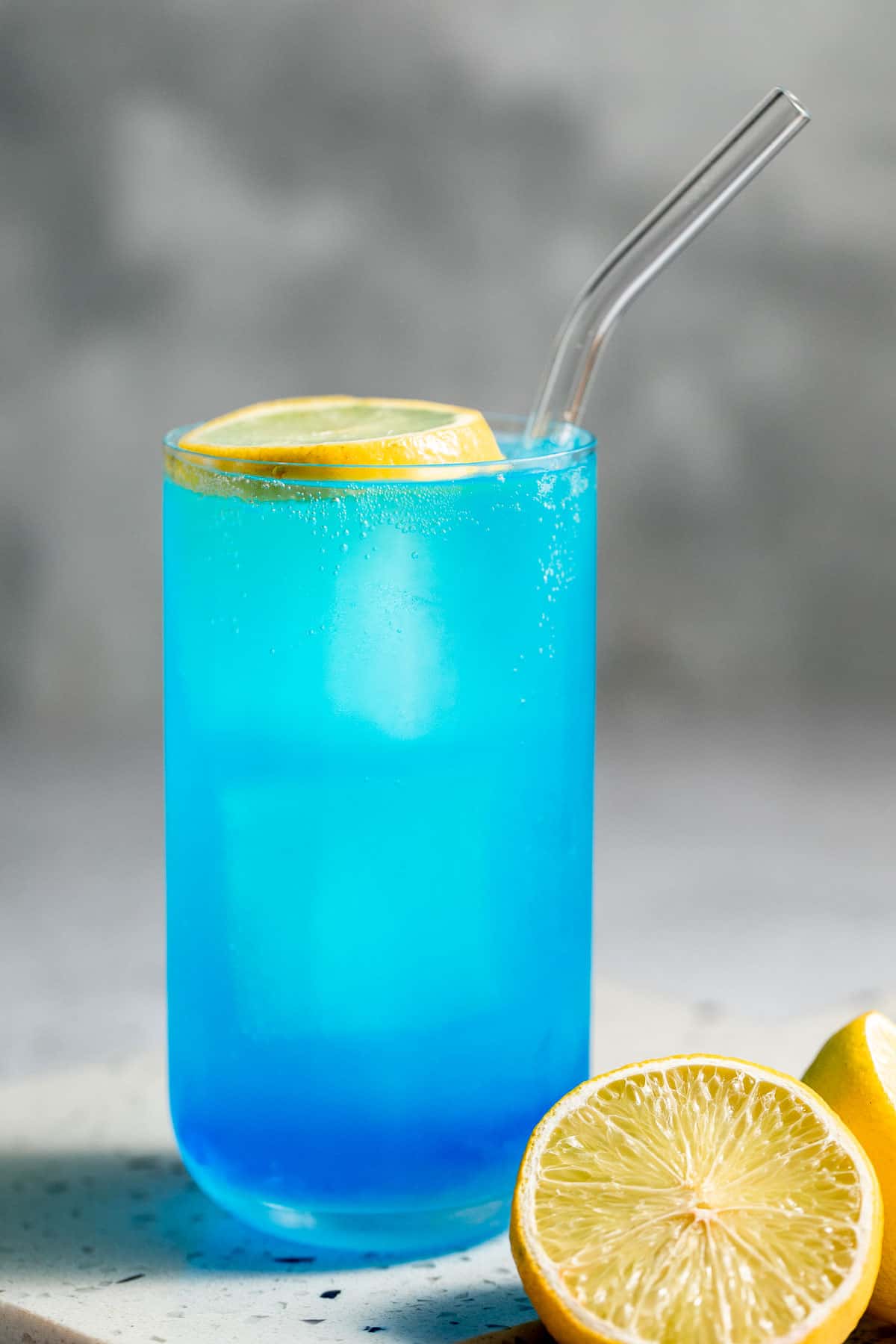 An aqua velva blue drink in tall glass with glass straw and lemon slice