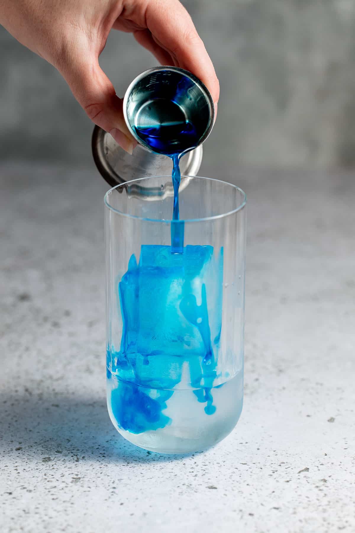 Blue curacao liqueur being poured into a tall glass with ice cubes.