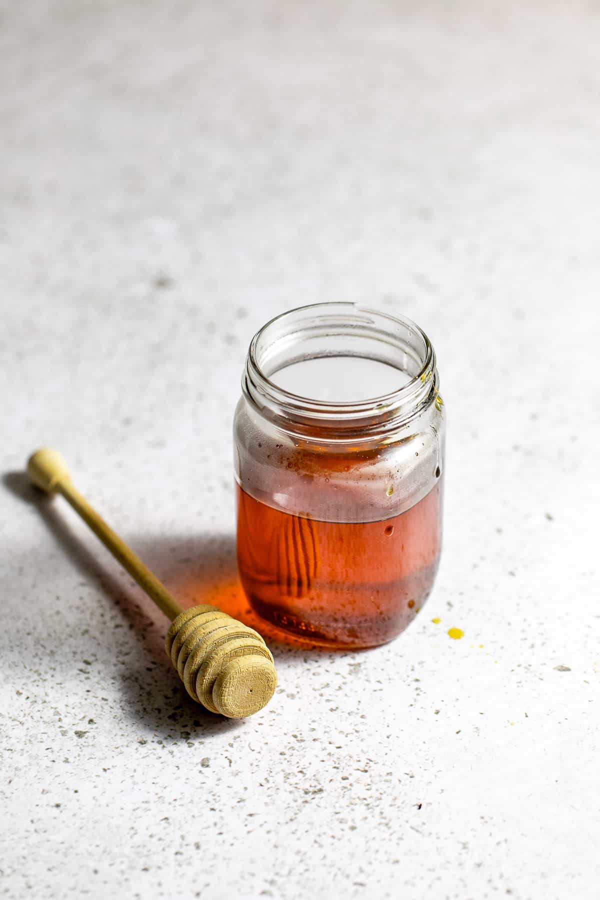 A jar of honey simple syrup sitting beside a honey dipper on a white table.