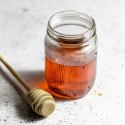 A jar of honey simple syrup sitting next to a honey dipper on a white table.