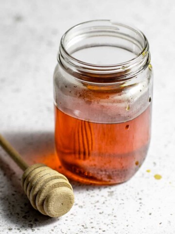 A jar of honey simple syrup sitting next to a honey dipper on a white table.