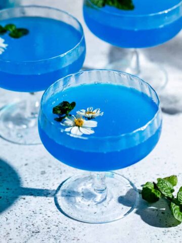 Three blue lagoon cocktails served in coupe glasses and garnished with fresh flowers.