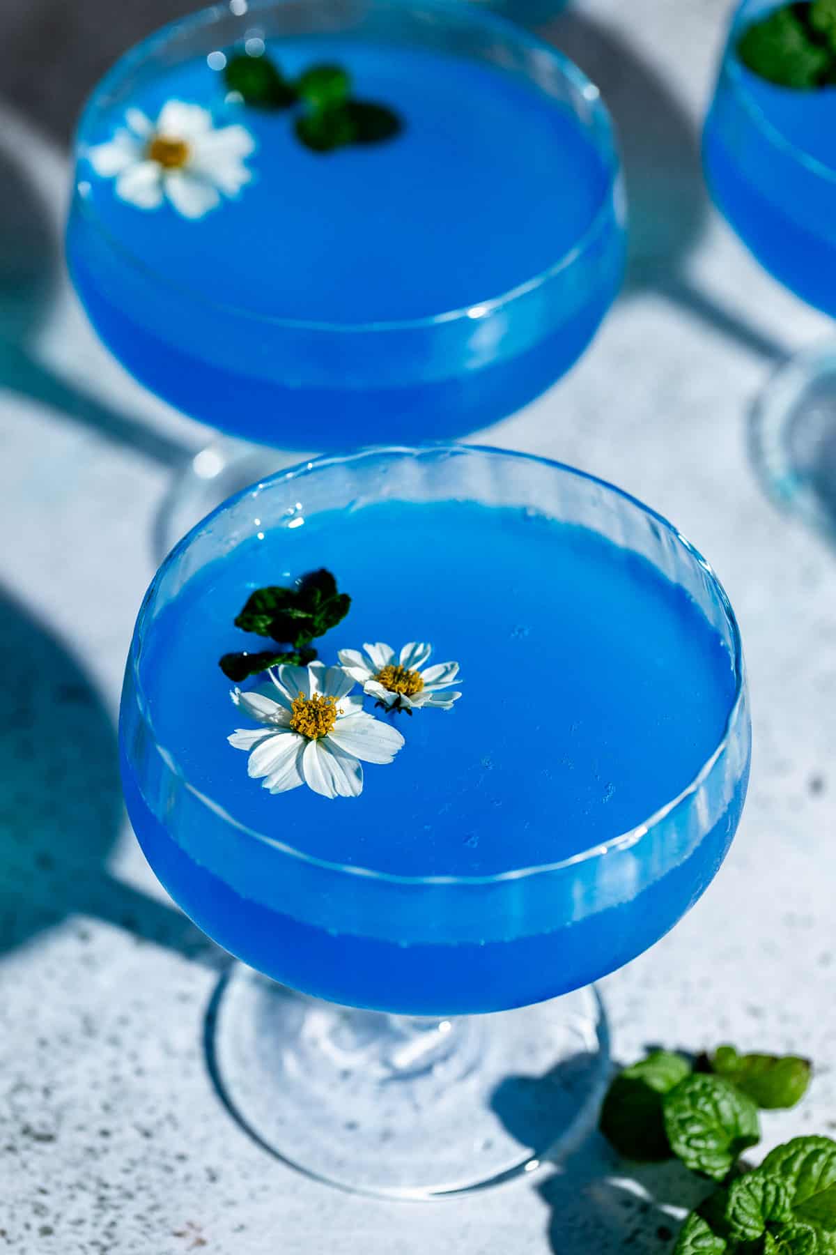 Two blue lagoon cocktails served in coupe glasses and garnished with fresh flowers.