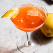 coupe glass with paper plane cocktail and garnished with lemon twist on grey background