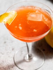 coupe glass with paper plane cocktail and garnished with lemon twist on grey background