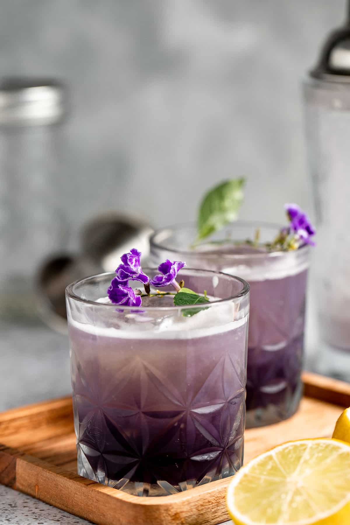 Two purple rain drinks topped with fresh purple flowers sitting on a wooden tray.