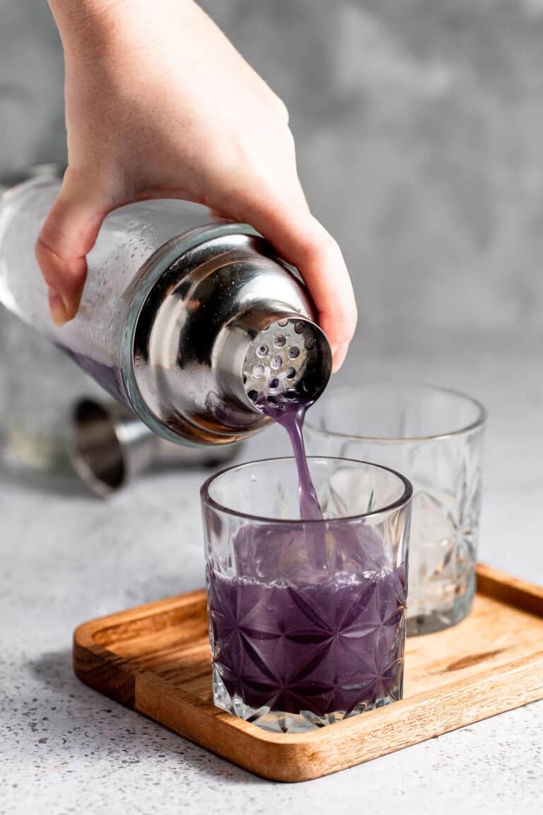 A purple cocktail being pour over ice in an old fashioned glass.