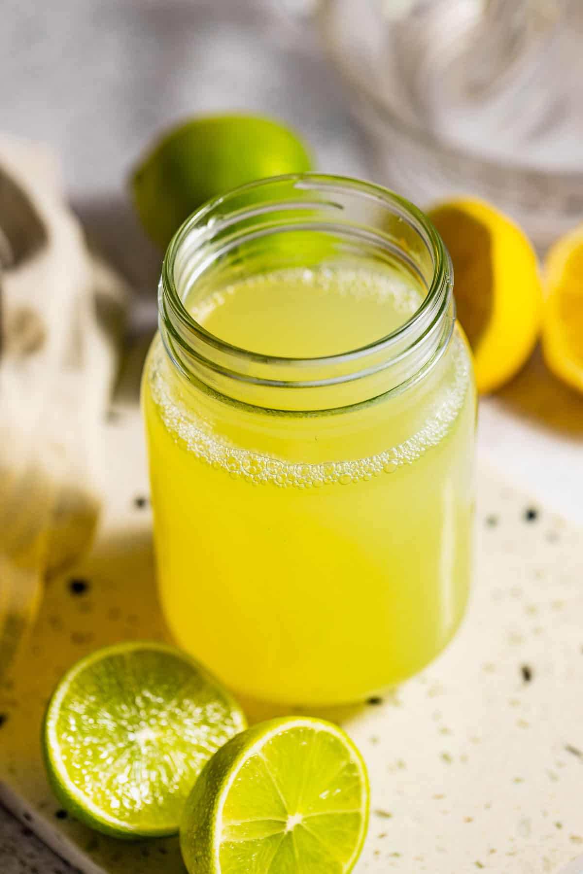 A jar of sweet and sour cocktail mix sitting on a counter next to sliced lemons and limes.