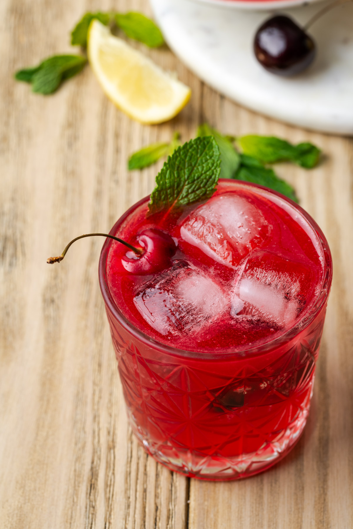 A rocks glass filled with cherry bourbon smash and topped with a mint leaf and fresh cherry.